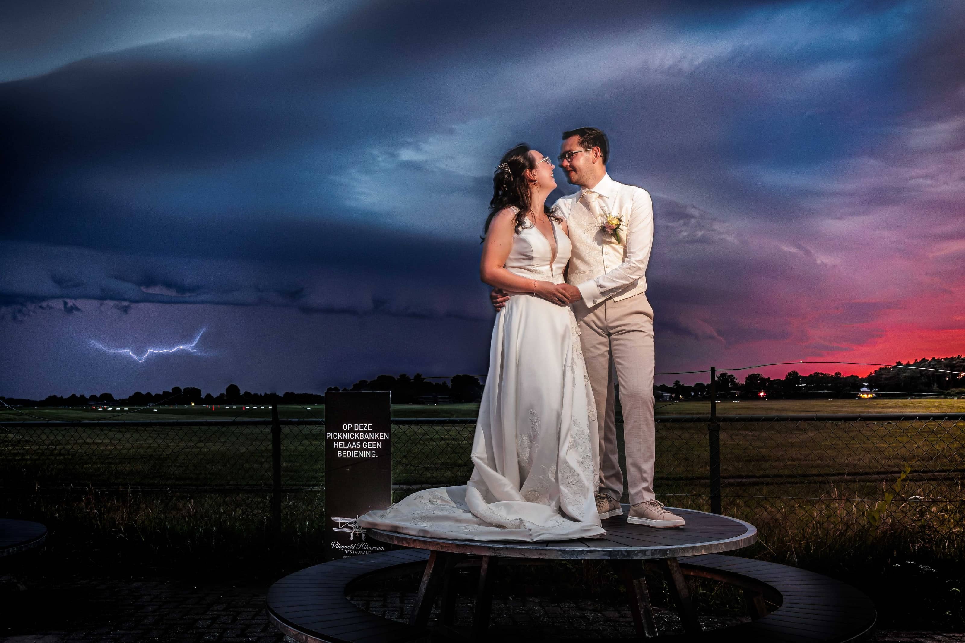 Newlyweds standing on a picnic table under a stormy sky with lightning and a red glow