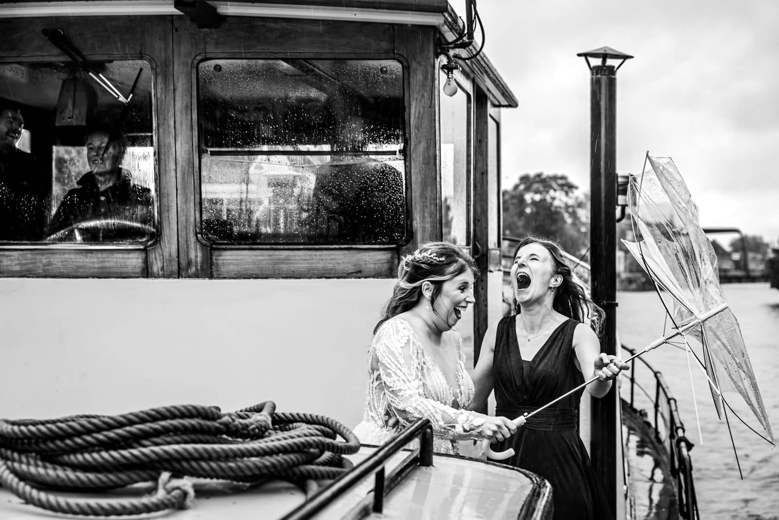 On a boat a friend roars with laughter beside the bride, holding a broken umbrella (black and white)
