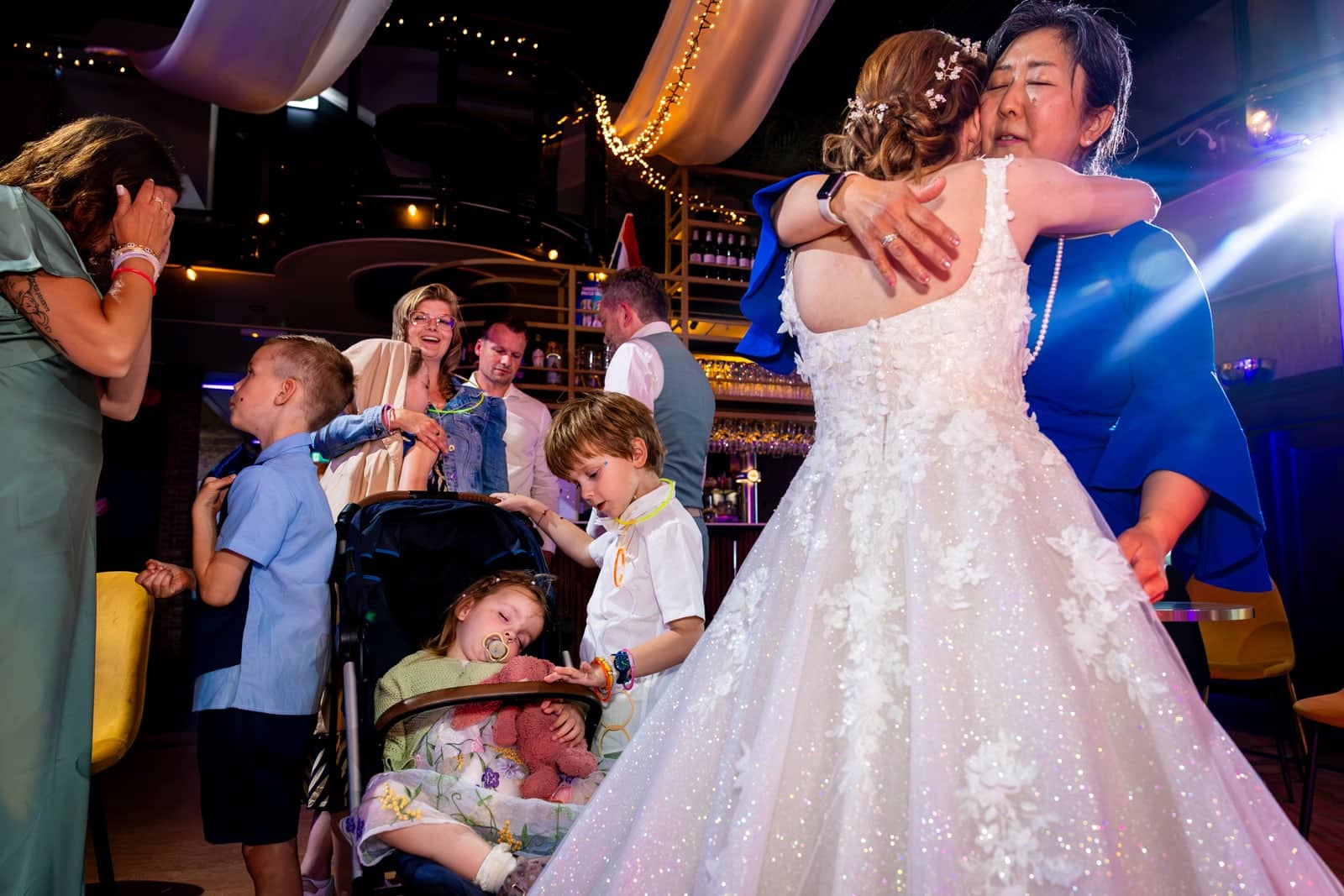 The bride hugs a guest on the dance floor, surrounded by children and a sleeping toddler