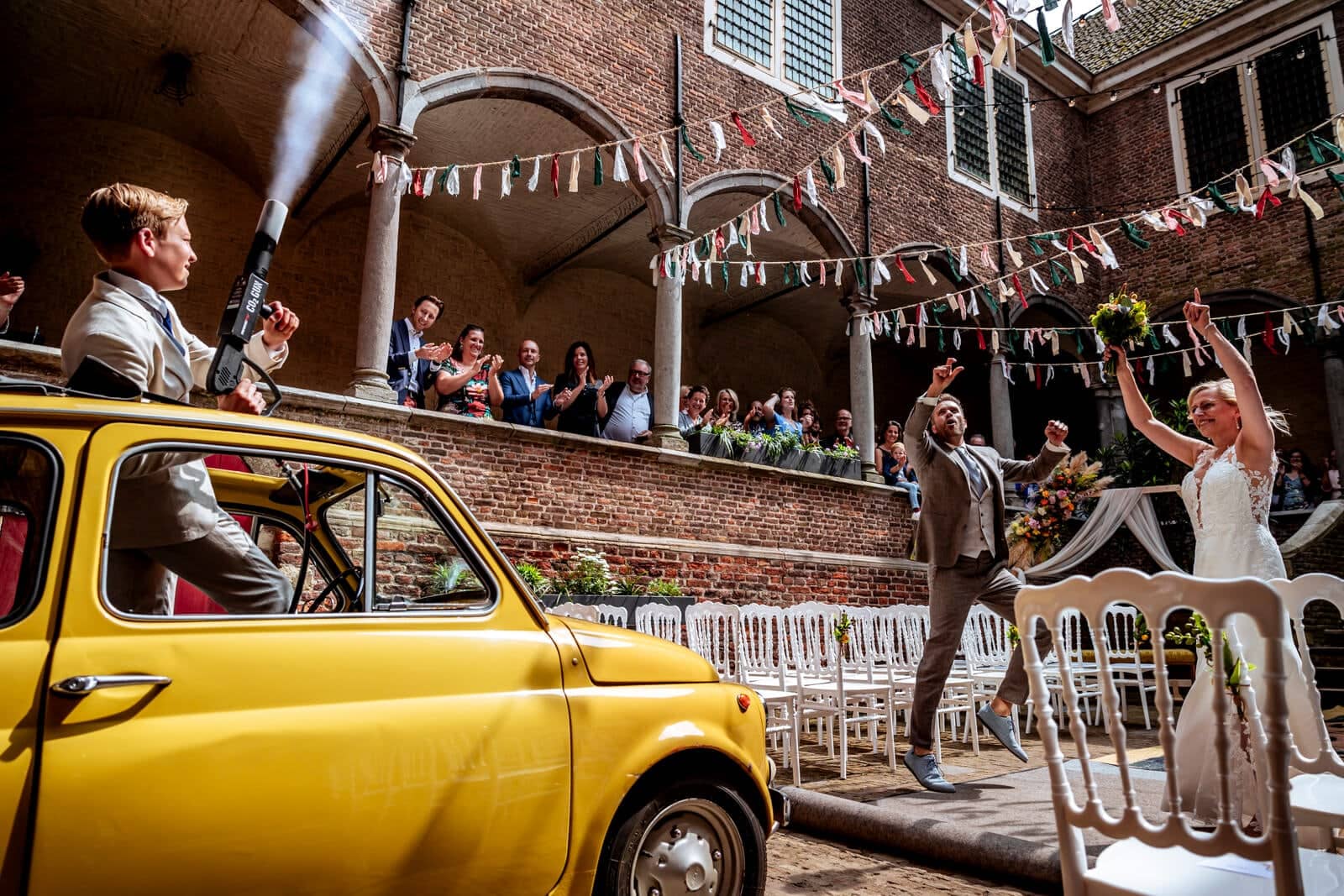 A guest sprays champagne from a yellow Fiat 500 as the couple cheer their way into the castle courtyard