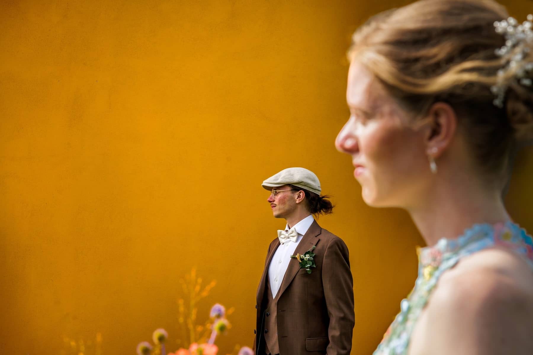 The groom in a flat cap against a yellow wall, the bride out of focus in the foreground