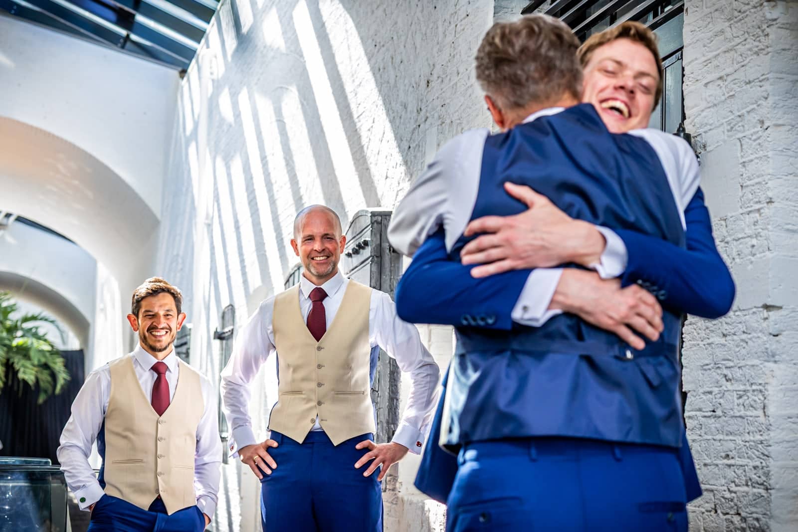 The groom hugs a friend while the best men in waistcoats laugh alongside