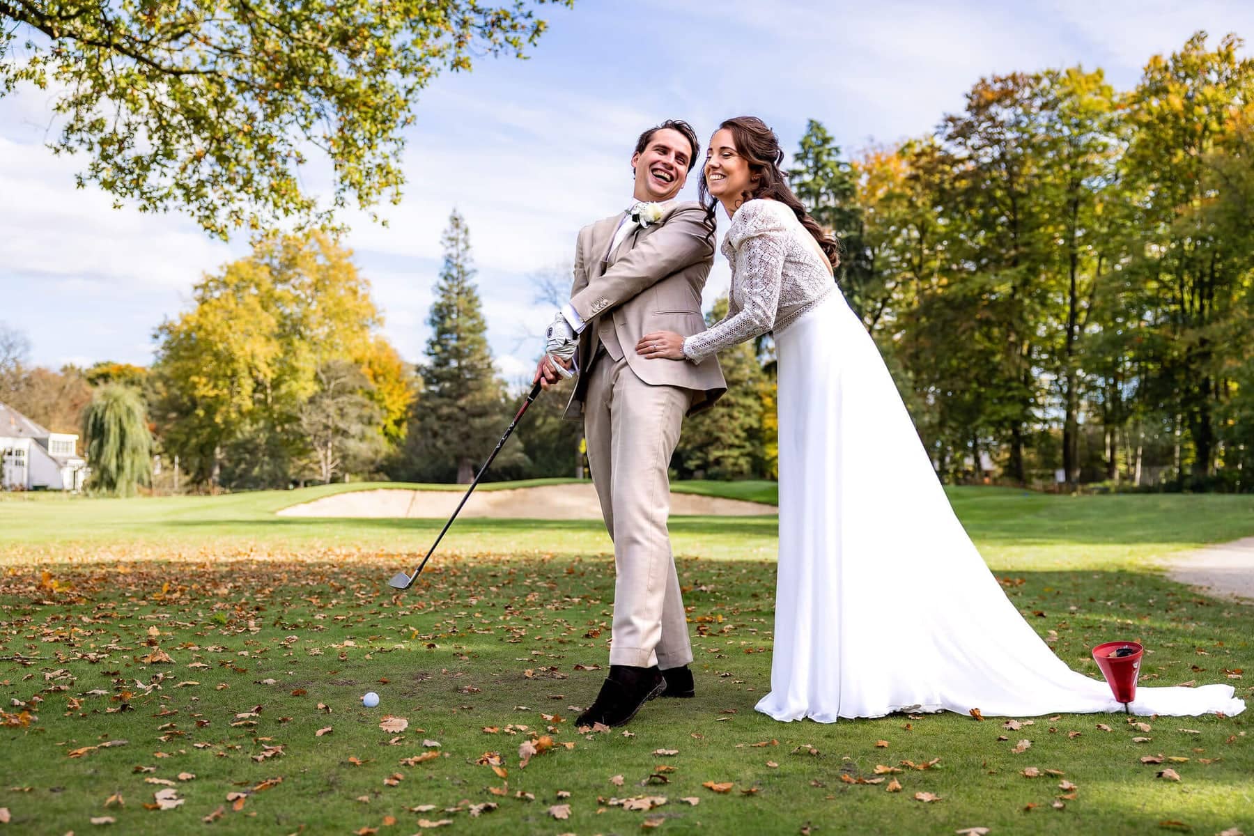 The groom tees off while the bride holds on laughing, on an autumn golf course