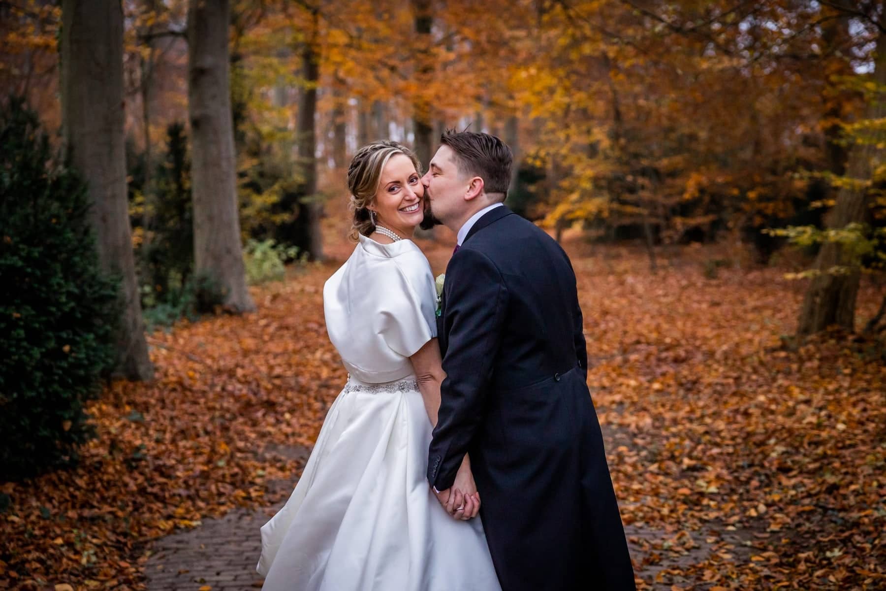 The groom kisses the bride on the cheek on a forest path covered in autumn leaves