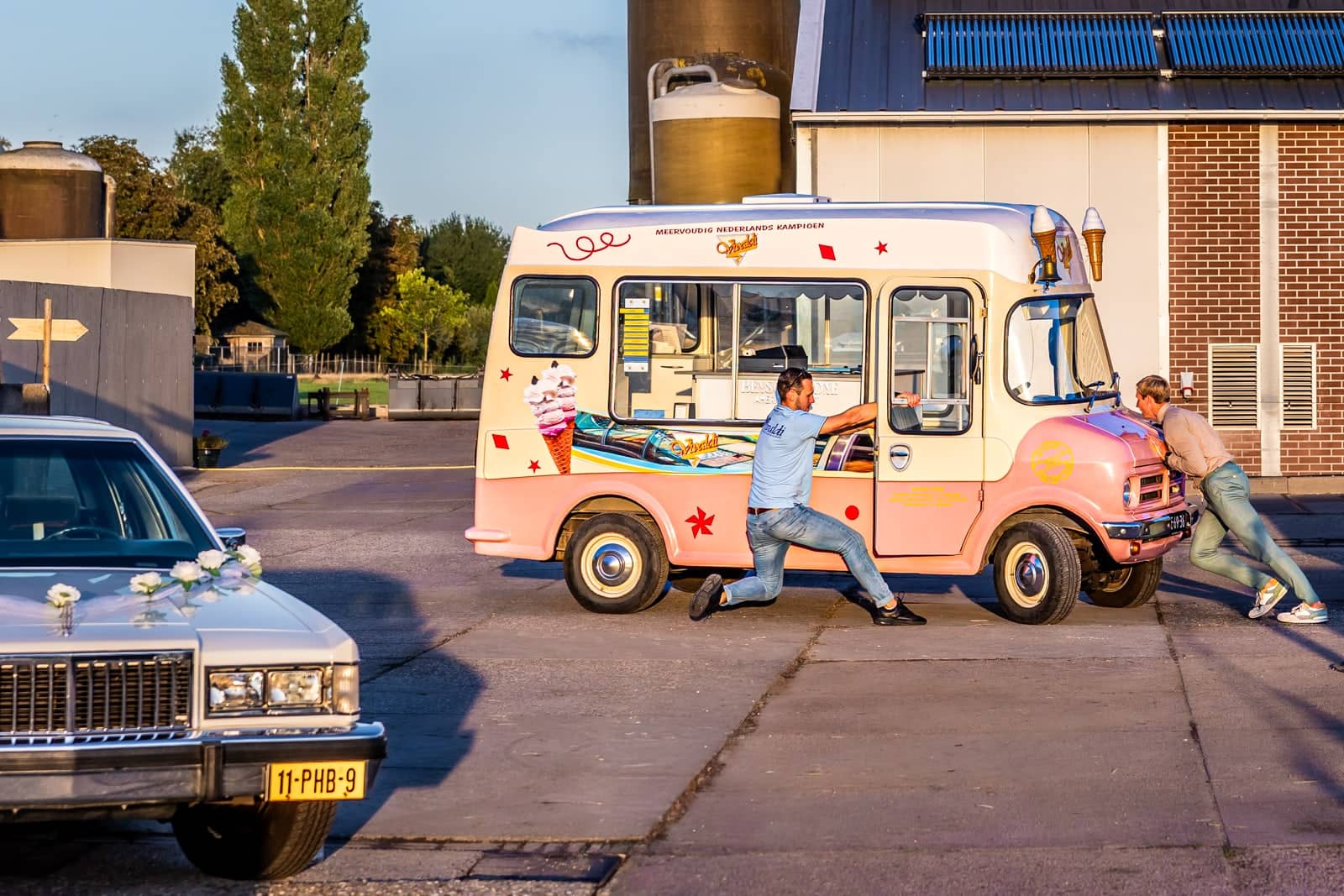 Two men push a pink ice-cream van next to a vintage white wedding car