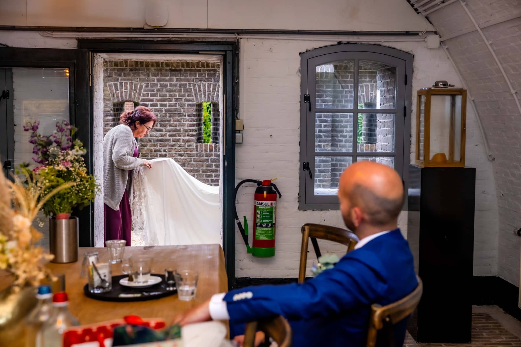 A woman spreads out the wedding dress while the groom waits at the table