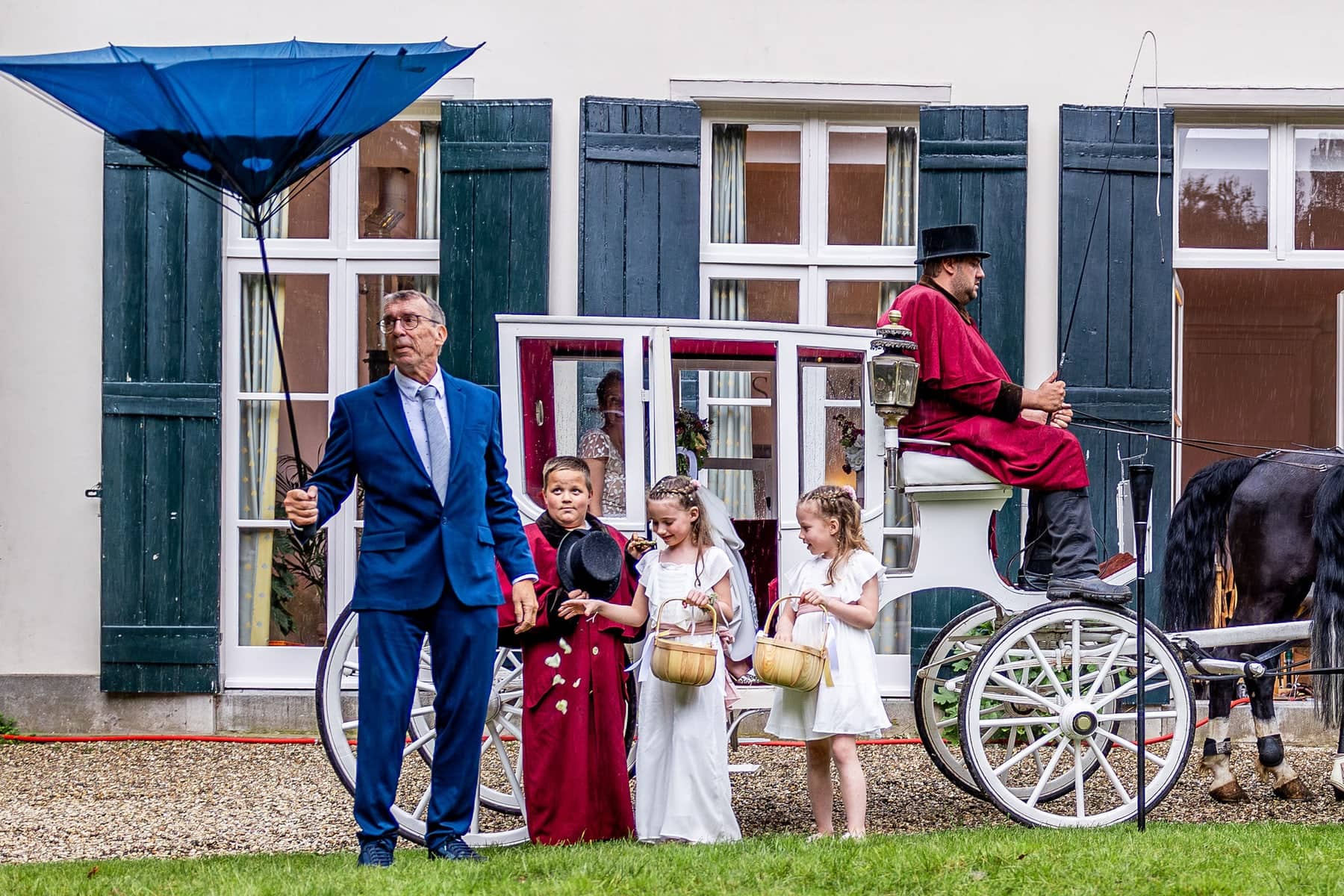 A man holds an umbrella blown inside out next to a white carriage with flower children