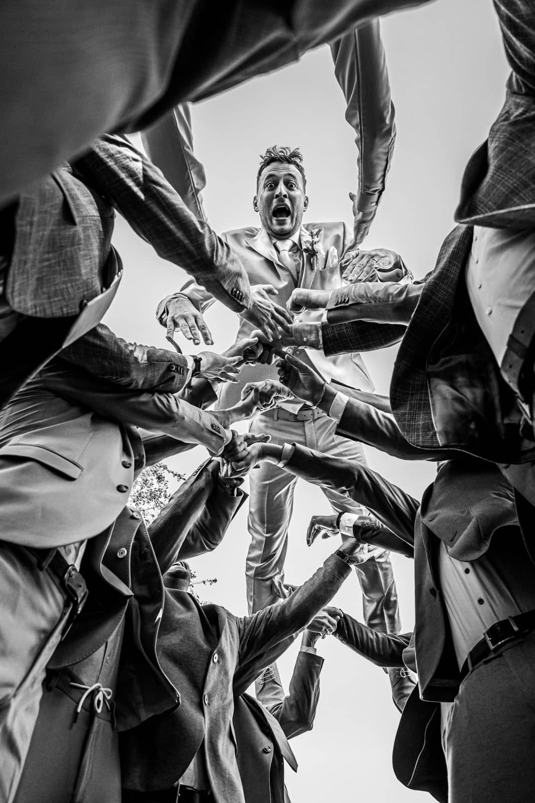 From below: friends raise their arms in a circle around the groom (black and white)