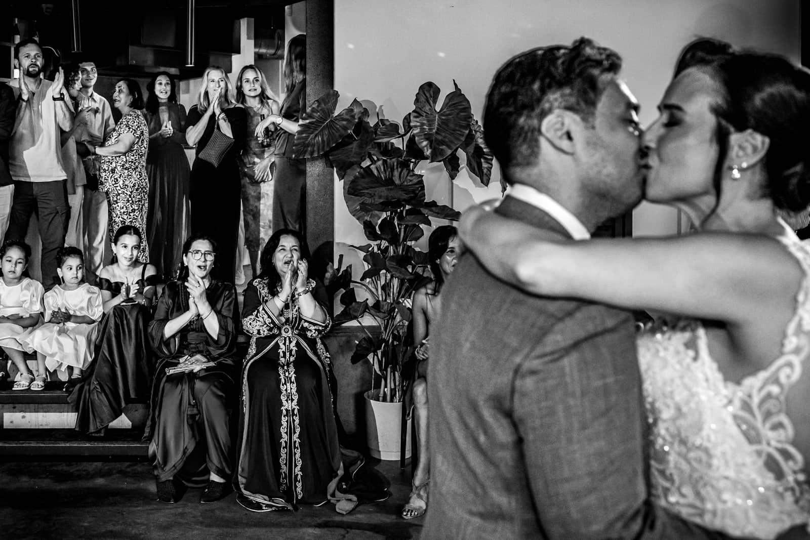 The couple kisses as guests cheer and clap (black and white)