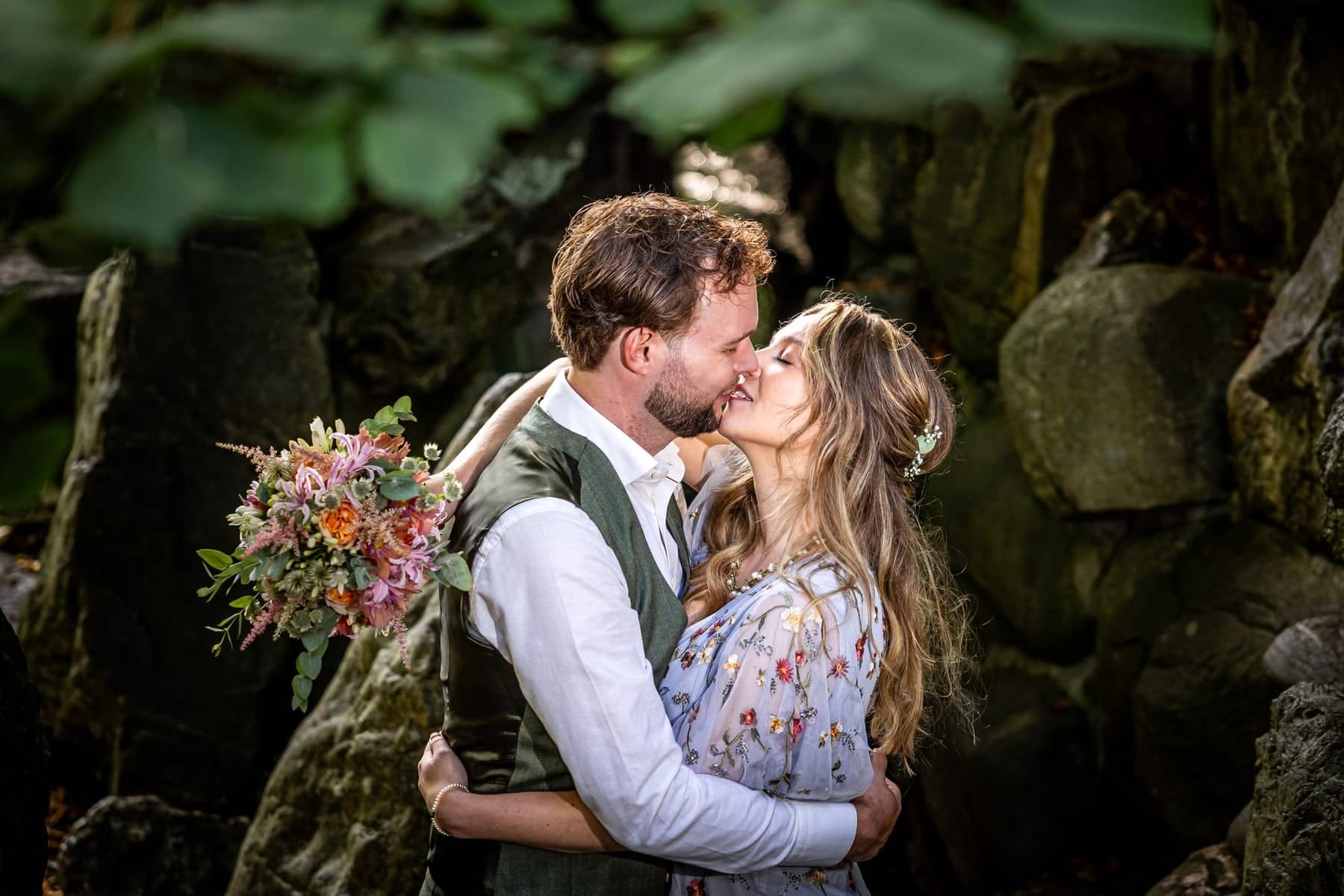 The couple kisses between large rocks in a wooded setting, the bride holding a colourful bouquet