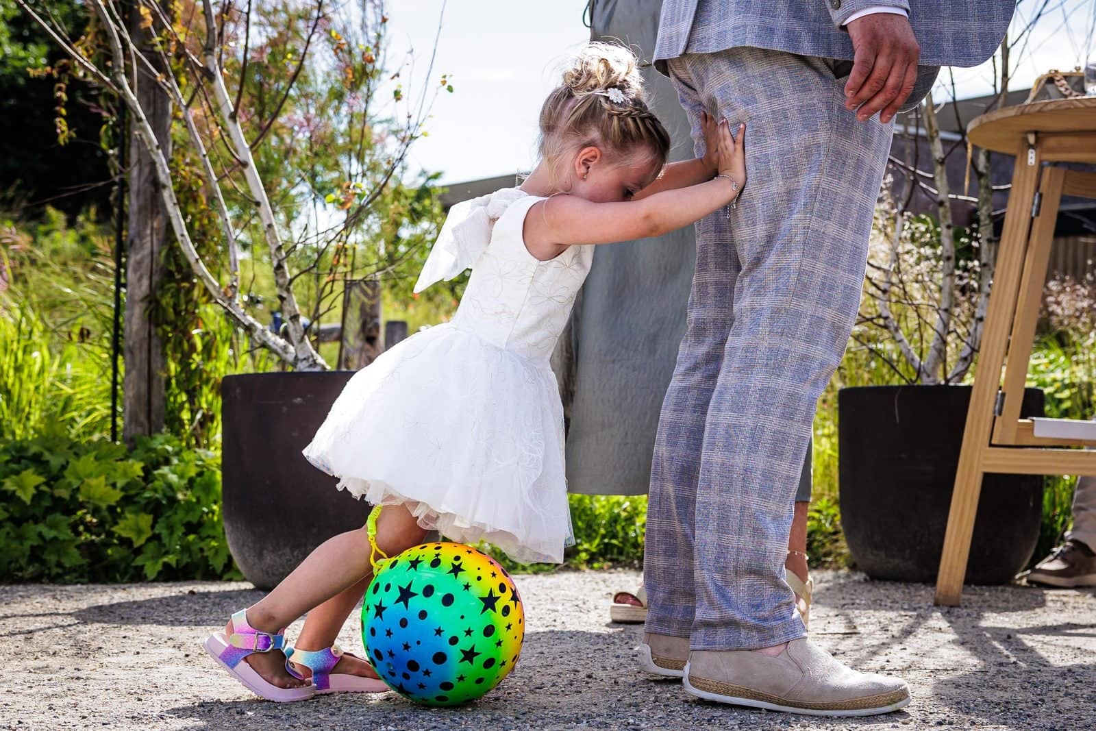 A girl in a white dress pushes against a guest’s leg with all her might, a colourful ball at her feet