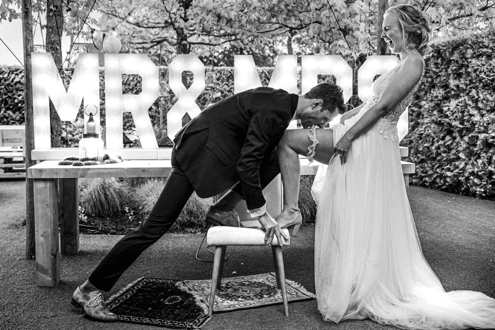 In front of large MR & MRS letters, the bride puts her foot on a stool for the garter (black and white)