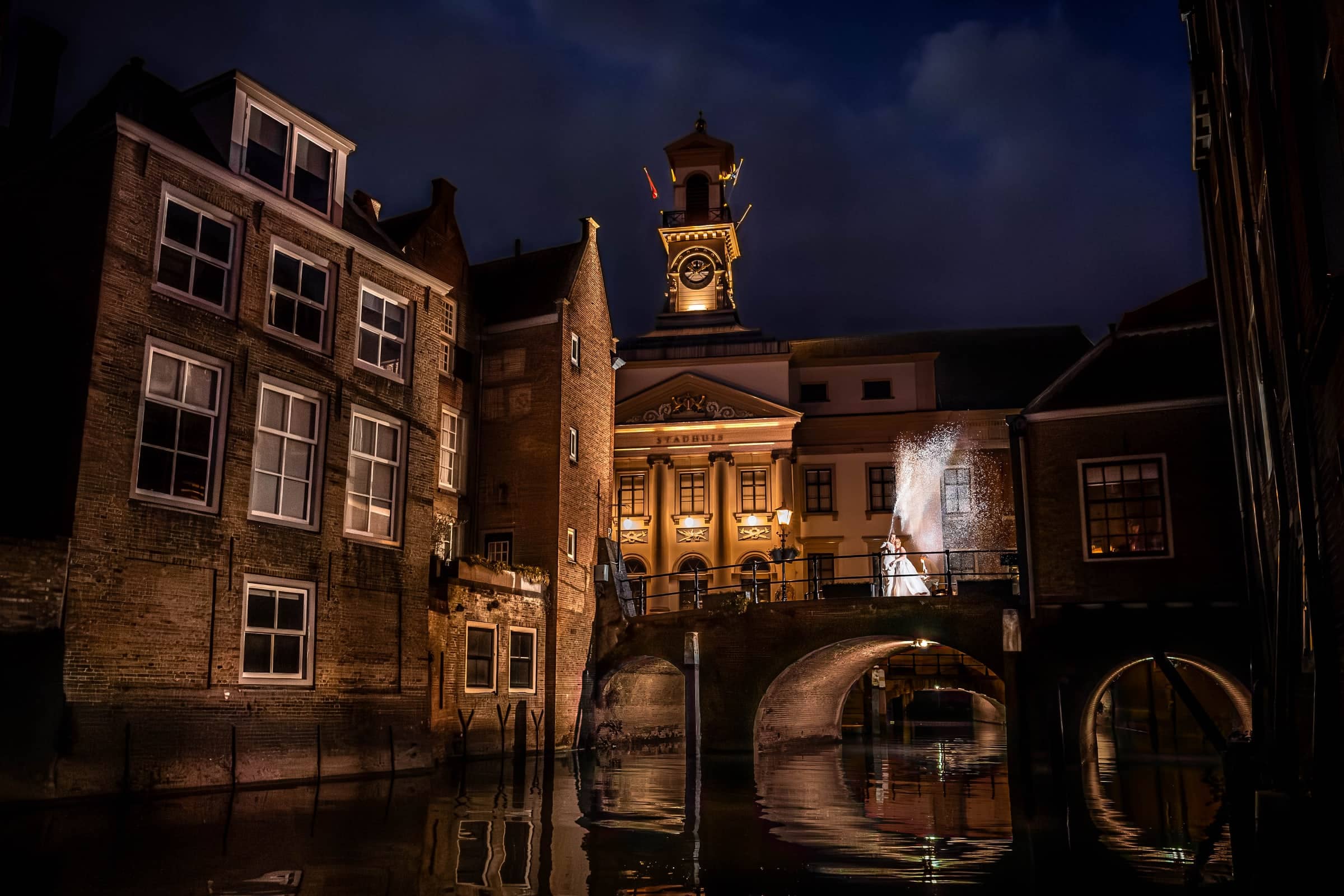Couple lit by flash by a canal bridge at night, between illuminated historic buildings