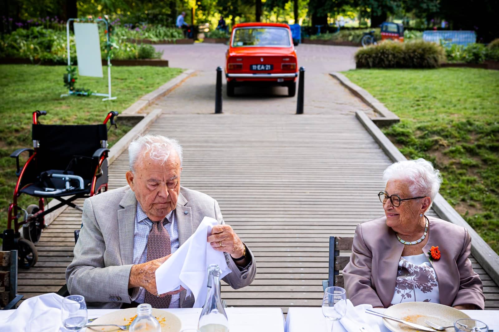 An elderly couple at the table, the man unfolding his napkin, a red vintage car in the background
