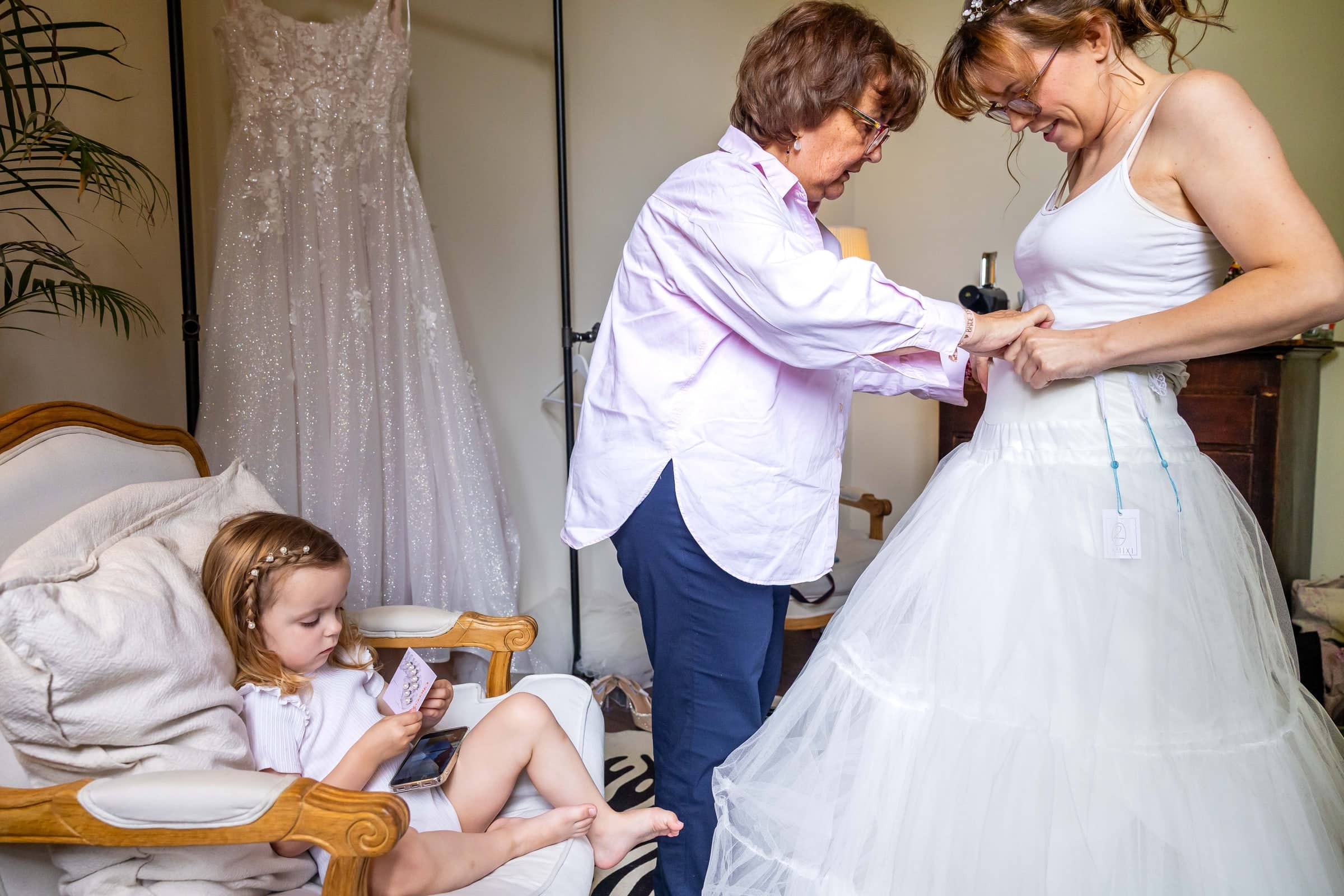 Mum helps the bride with her petticoat while a little girl reads in a chair