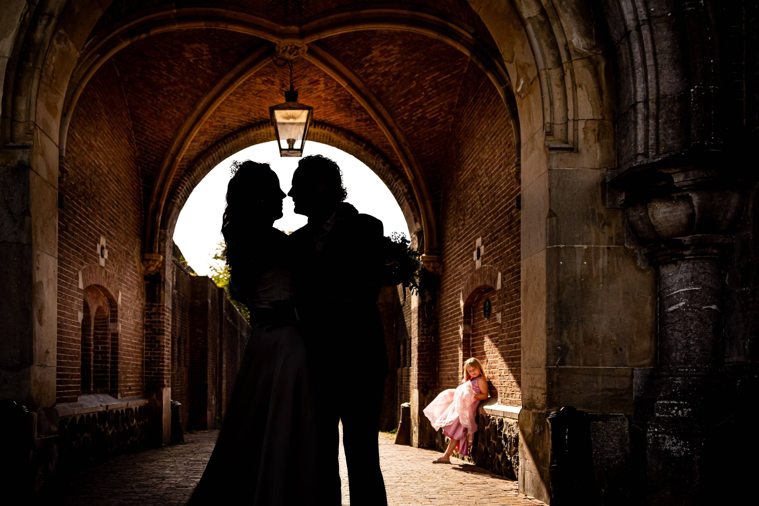 Silhouette of a couple kissing under an archway, with a flower girl waiting in the background