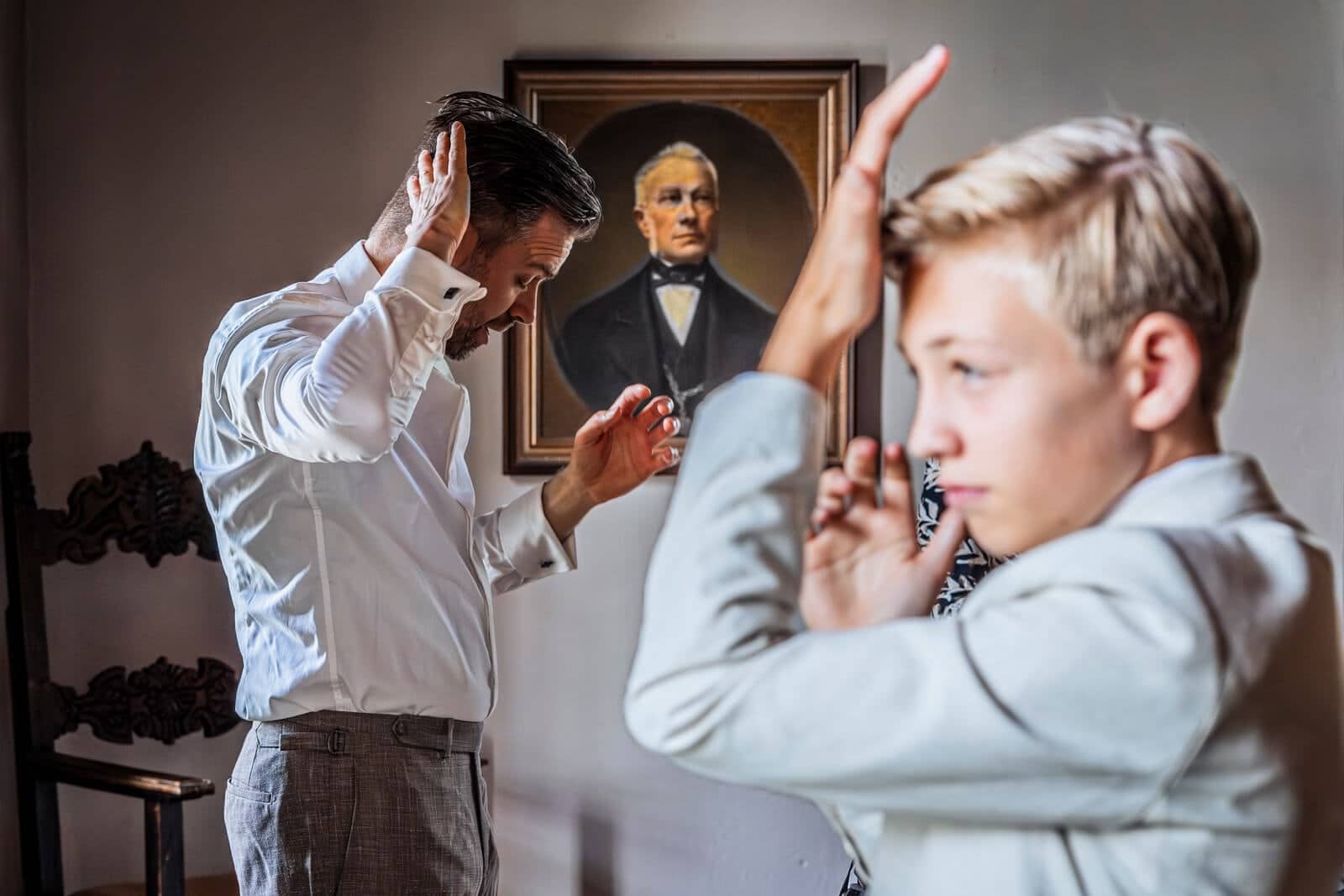 A man and a boy with a hand to their foreheads, in front of an old portrait on the wall