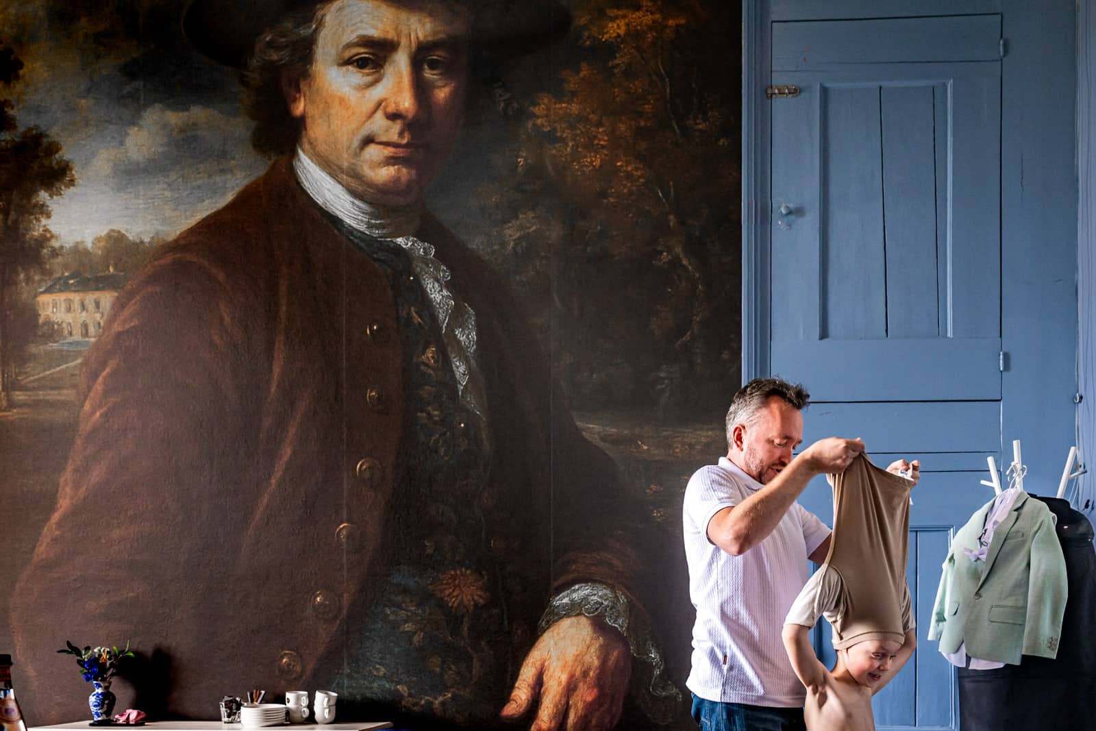 Beneath a large painted portrait, a man helps a little boy get dressed