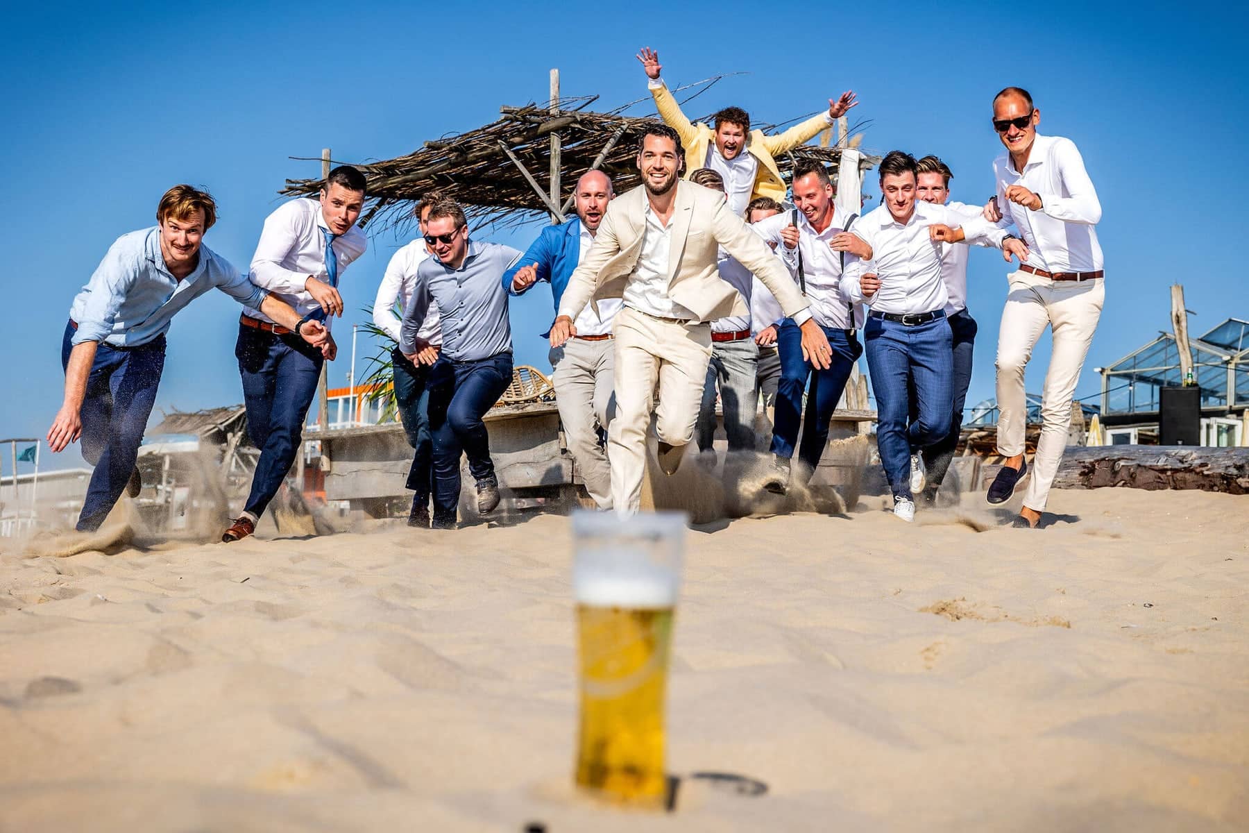 The groom and his friends sprint across the beach towards a glass of beer
