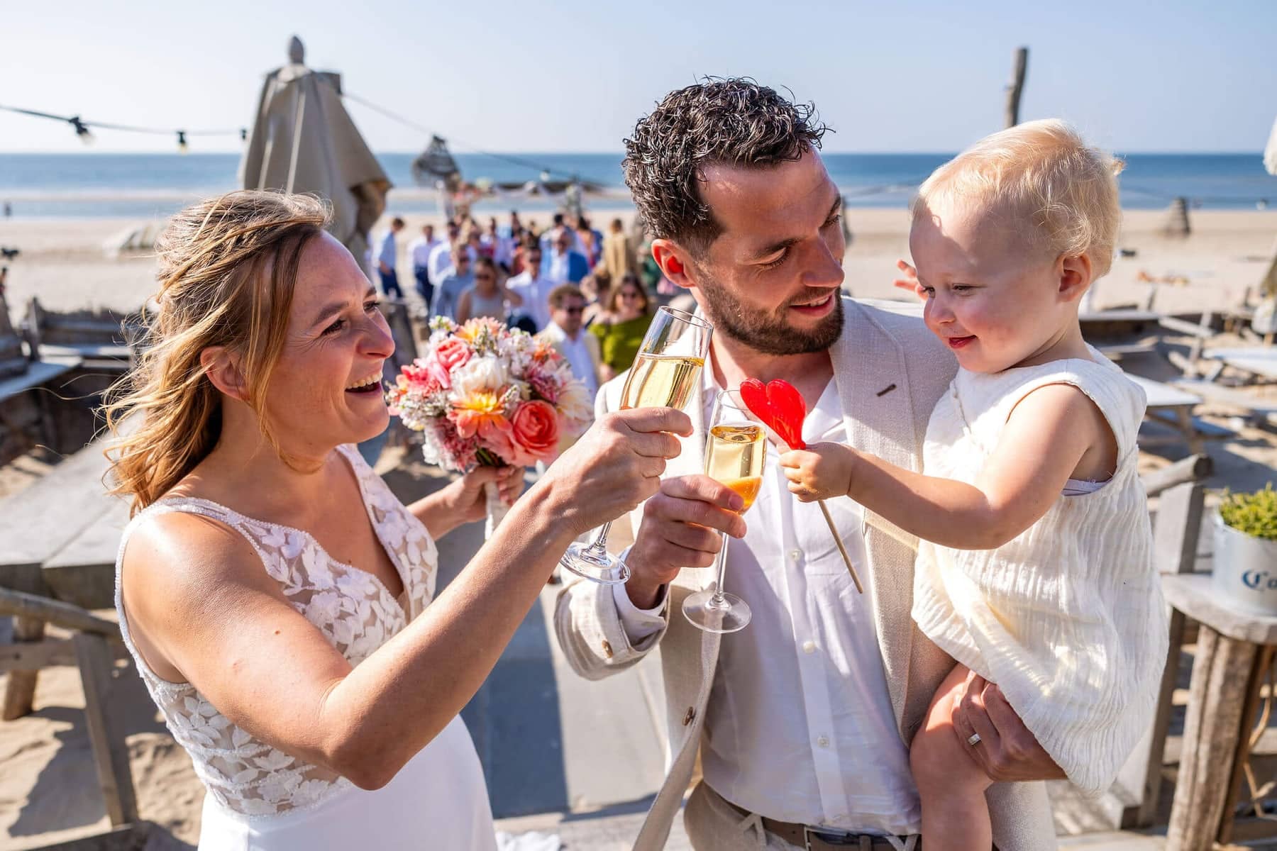 The couple toast with champagne on the beach, the groom holding a laughing toddler