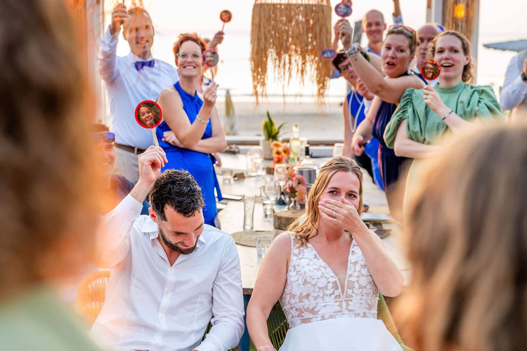 The bride covers her mouth as guests at the beach club hold up photos on sticks