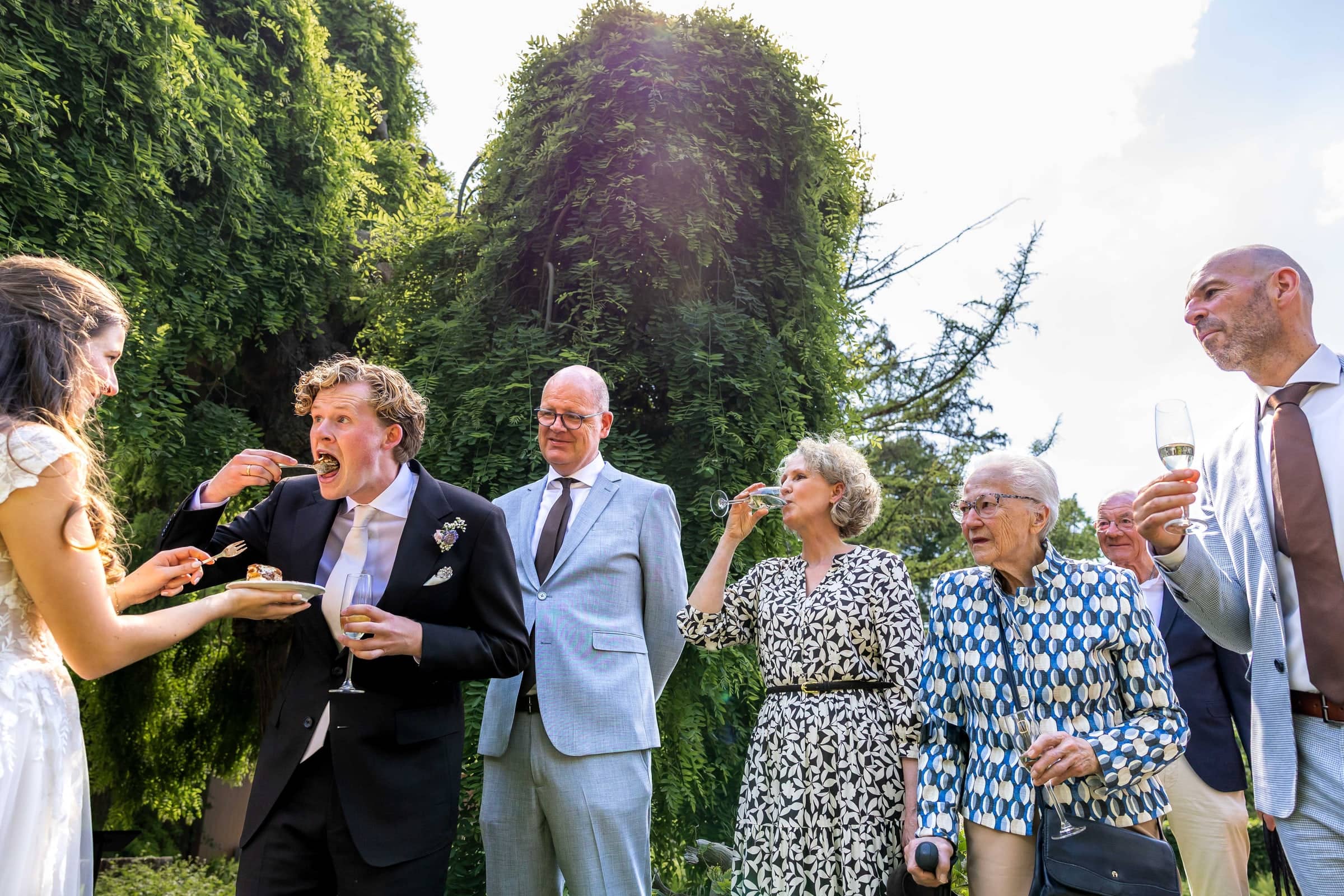 The bride feeds the groom a big bite of cake while the family laughs