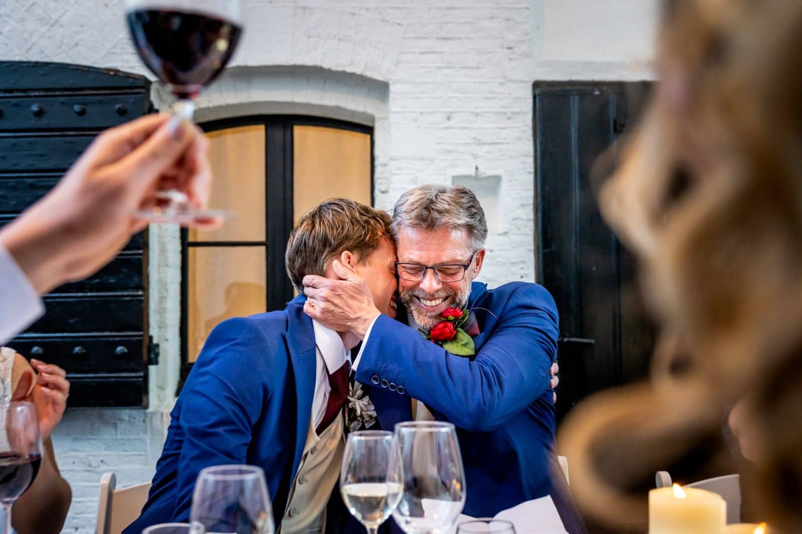 The groom hugs his father during a toast, glasses of red wine raised