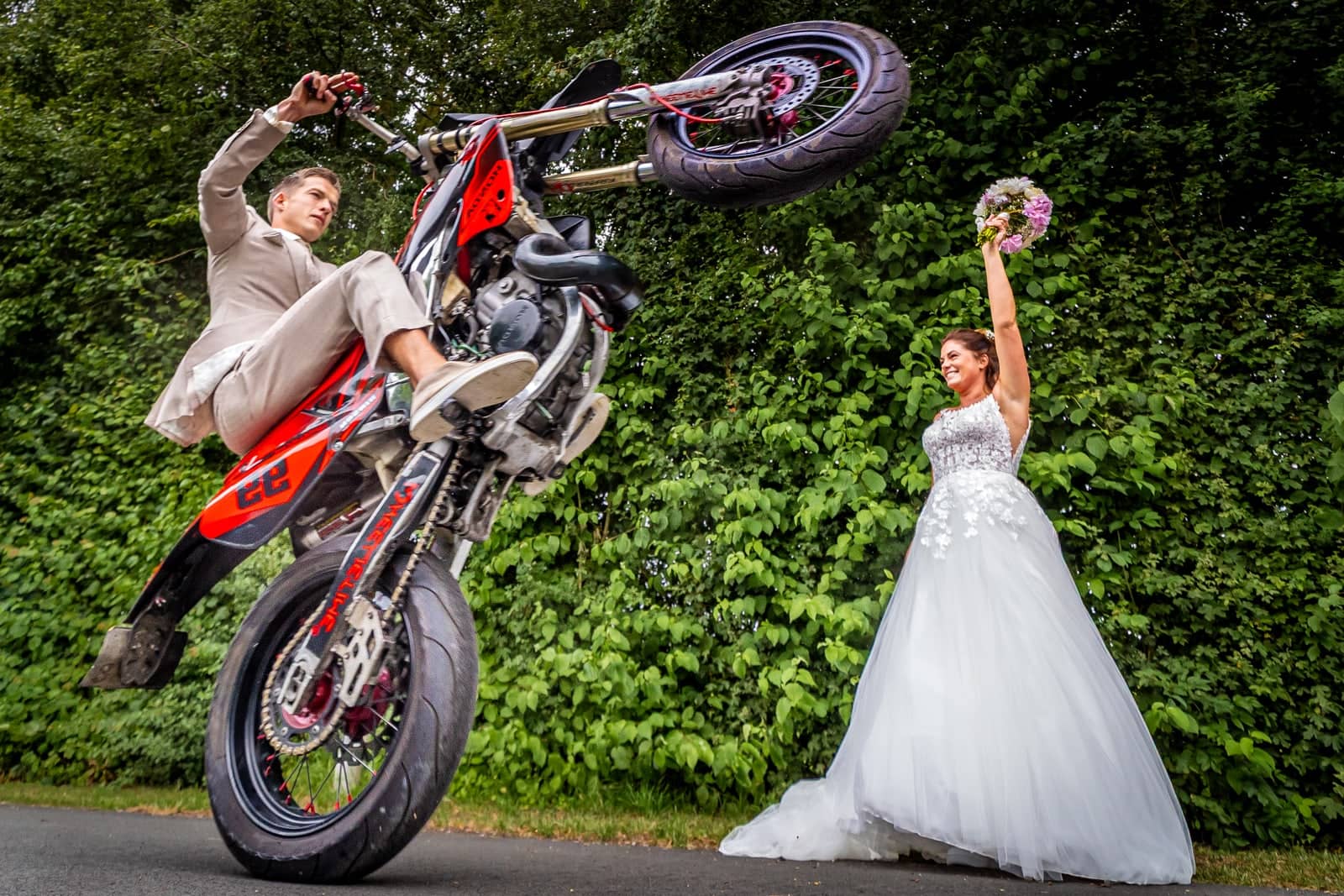 The groom pulls a wheelie on his motorbike as the bride cheers with her bouquet in the air