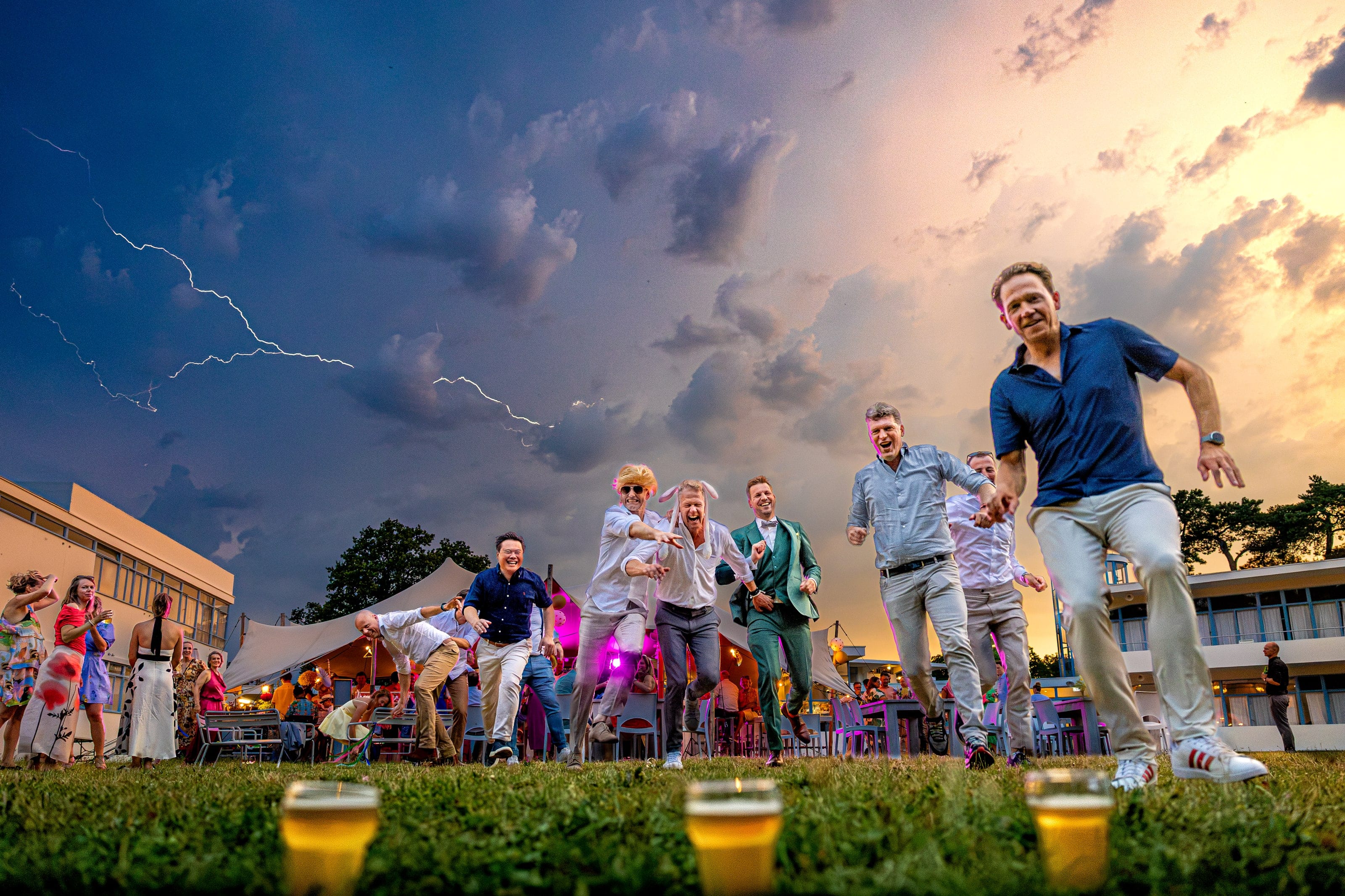 Wedding guests sprint laughing towards their beers on the lawn while lightning strikes behind them
