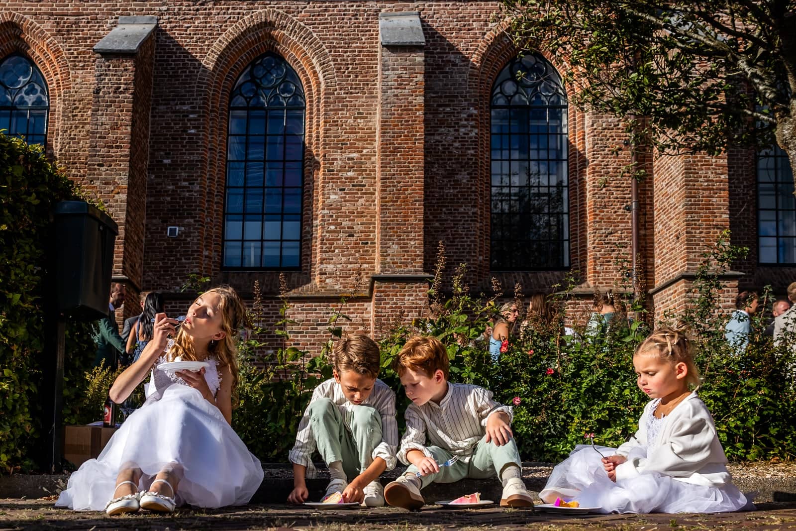 Page boys and flower girls sit on the grass in front of the church eating cake