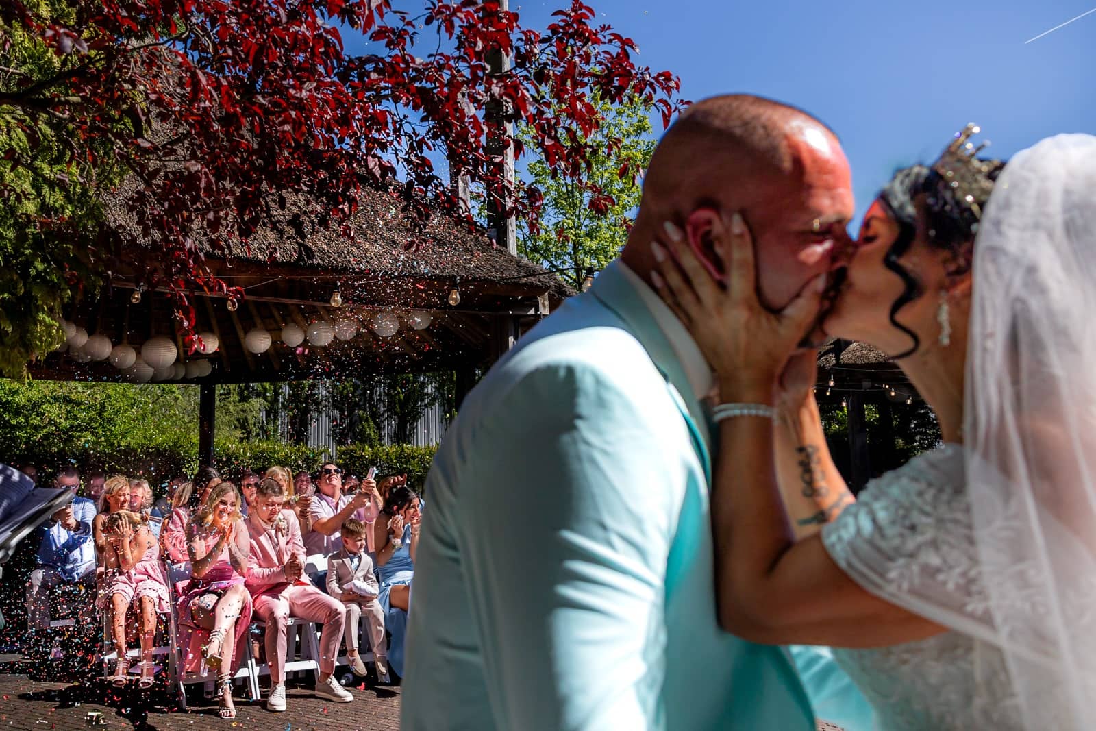 Bruidspaar kust bij de ceremonie in een zonnige tuin, kinderen op de eerste rij kijken toe