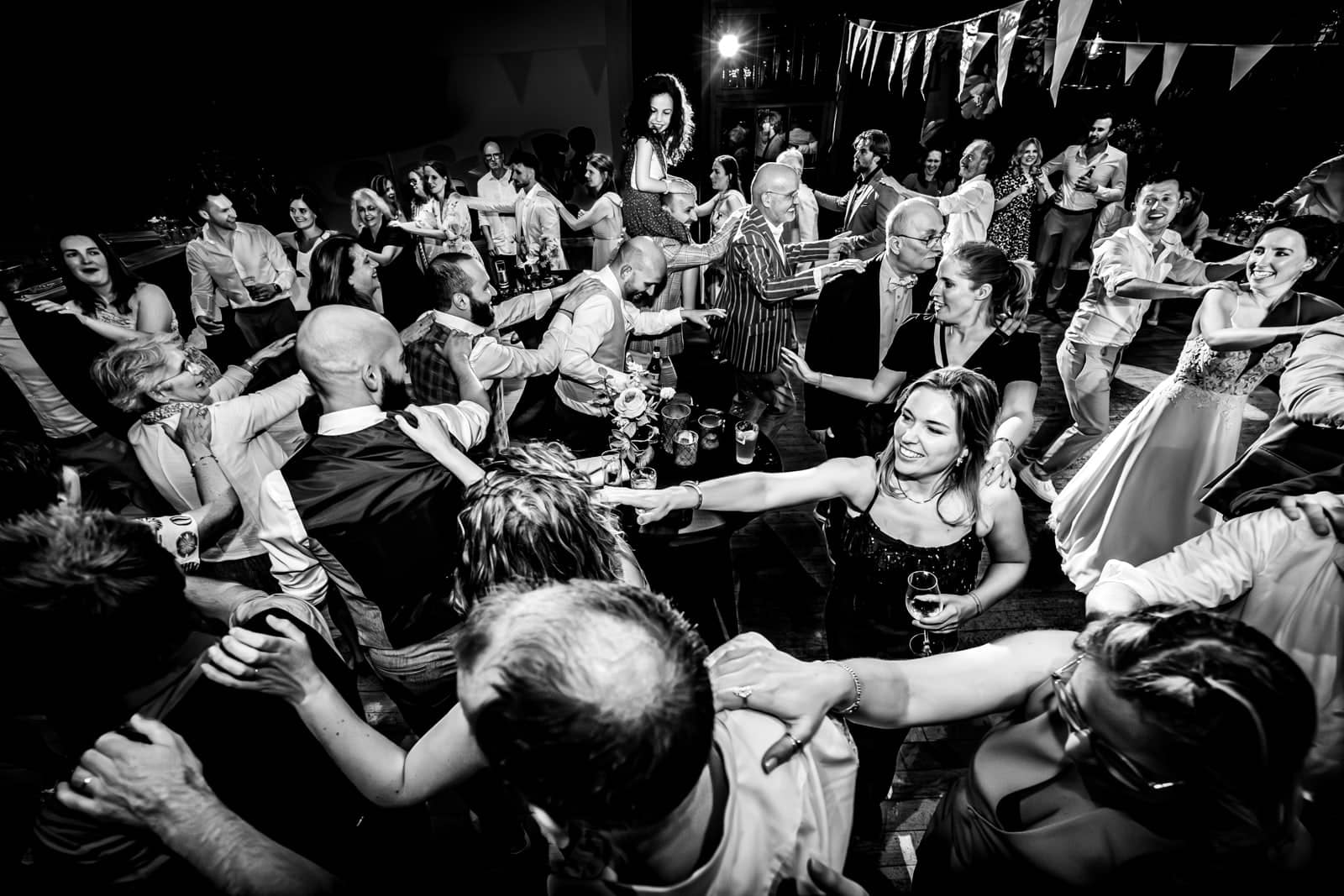 The whole room joins a conga line, with a laughing girl on someone’s shoulders in the middle (black and white)