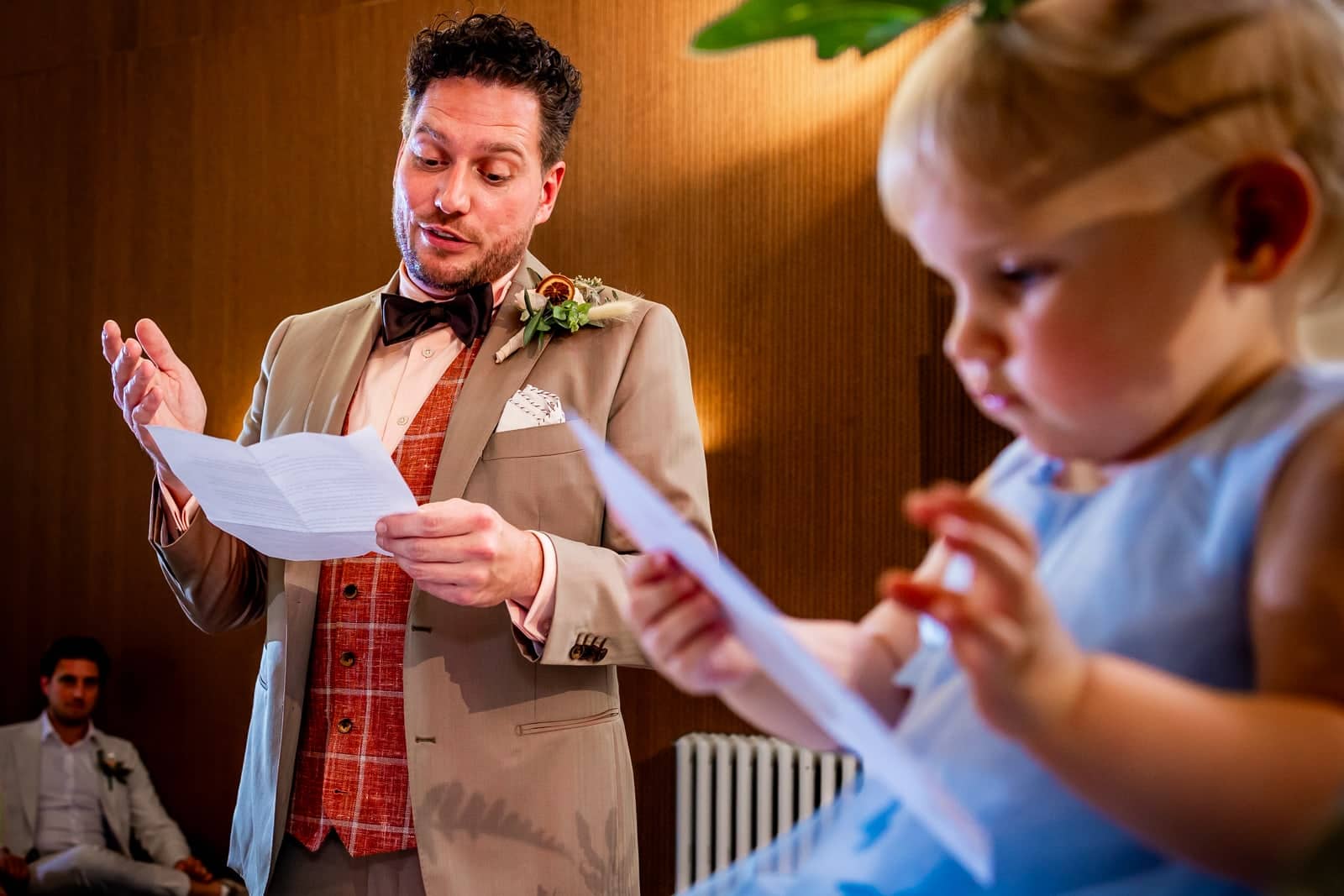 De bruidegom houdt een speech terwijl een baby op de voorgrond met een papiertje speelt